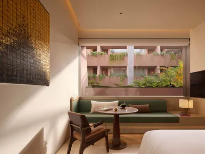 Modern hotel room featuring a green daybed, walnut armchair, round wooden table, large picture window overlooking a pink courtyard with hanging plants, and a textured gold artwork on the wall.