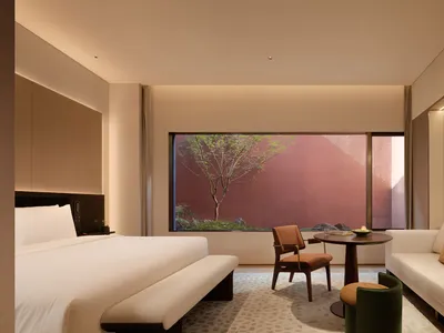 Modern luxury hotel room featuring a large bed with white linens, a white sofa, wooden armchair, round coffee table, and a large window overlooking a private Japanese-style garden with a red wall and small tree.