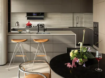 Modern minimalist kitchen with grey matte cabinetry, a central island with two leather bar stools, and a round black marble dining table with a floral arrangement in the foreground.