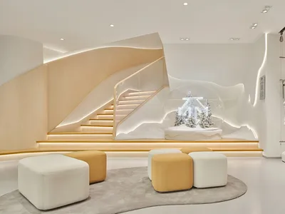 Modern minimalist retail interior with a large illuminated snow globe display, curved oak staircase with integrated lighting, and modular white and mustard ottomans on a grey rug.
