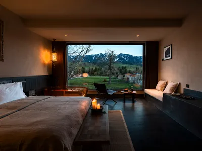 Modern bedroom interior featuring a large bed with taupe linen, a freestanding copper bathtub, built-in window seat, and dramatic mountain landscape visible through floor-to-ceiling windows.
