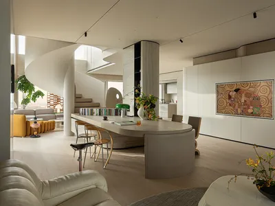 Modern open-plan living room featuring a dramatic white spiral staircase, curved light oak kitchen island with bar stools, cream leather sofa, and large Klimt-style artwork on the wall.