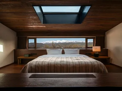 Modern minimalist bedroom with dark wood ceiling, large skylight, striped bedding, and panoramic snow-capped mountain view through wide windows.