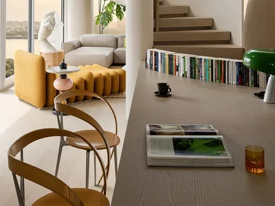 Modern minimalist living room featuring a dramatic curved oak spiral staircase, mustard yellow sofa, wooden chairs, and large windows with city view