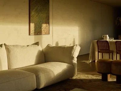 Minimalist living room featuring a cream bouclé sectional sofa, marble coffee table with art books, textured rug, and warm sunlight creating dramatic shadows on a textured wall with a landscape painting.