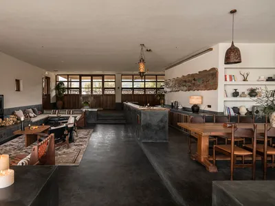 Modern open-plan living, dining and kitchen space with dark concrete floors, wooden dining table, built-in sofa, fireplace, and large windows overlooking nature.
