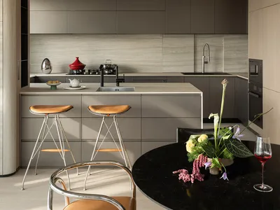 Modern minimalist kitchen with grey matte cabinetry, central island with two leather bar stools, and a round black marble dining table set with a vibrant floral arrangement and red wine.