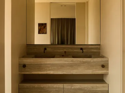 Modern minimalist bathroom with a long floating travertine double vanity, two black wall-mounted faucets, large frameless mirror, and warm beige tiled floor