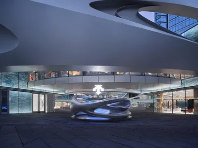 Modern retail atrium featuring a large flowing concrete sculpture on a circular platform, dramatic curved architectural ceiling, and glass storefronts with blue water-like wall panels.
