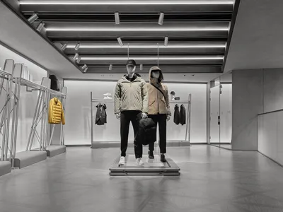 Modern minimalist menswear retail store interior with raw concrete walls, geometric floor tiles, linear LED ceiling lights, and two mannequins dressed in contemporary jackets displayed on a central metal platform.