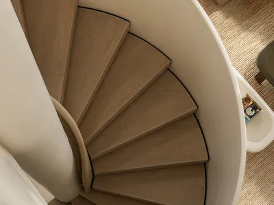 Top-down view of a minimalist spiral staircase made with light oak treads and a smooth white curved central column, adjacent to a woven natural-fiber rug and neutral sofa.