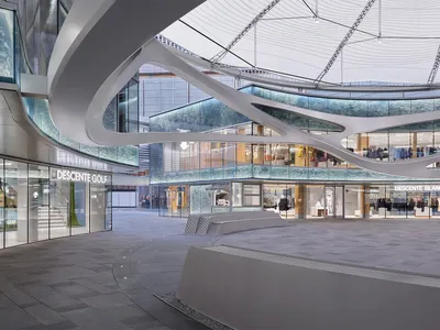 Modern multi-level retail atrium featuring dramatic sweeping white sculptural ceiling forms, glass storefronts for Descente Golf and Descente Blanc, and grey stone flooring.