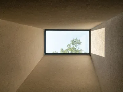 極簡室內角落，米白色質感灰泥牆面與天花板，大型橫向窗戶可見綠色樹梢與明亮天空，強烈陽光在右側牆面投射出矩形光影。
