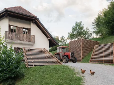 White alpine farmhouse with wooden balcony, large wooden pallet staircase, red vintage tractor, and two chickens on a gravel path with modern wooden slat structures on a grassy hill.