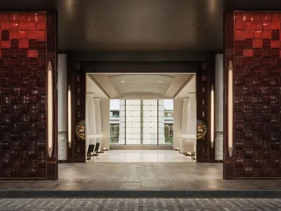 Luxurious hotel lobby entrance with large red tiled walls, vertical light sconces, white columns, and a view through to a bright courtyard.
