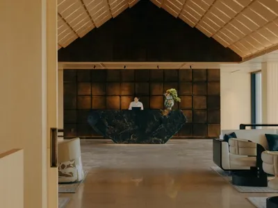 Modern luxury hotel lobby viewed from an entrance, featuring a black marble reception desk, warm wood-paneled ceiling with geometric folds, and contemporary seating area.