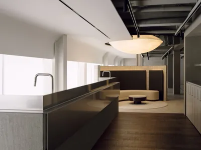 Modern brutalist commercial lounge featuring a long concrete counter with stainless steel faucet, dark wood flooring, a curved beige banquette, round coffee table, and a large glowing pendant lamp under an exposed ceiling.