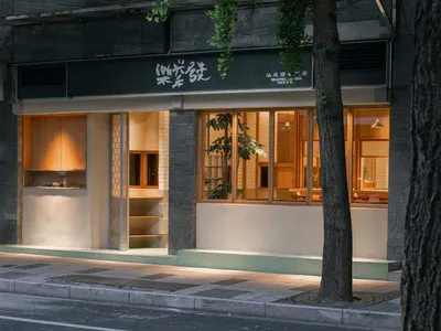 汕尾糯米店街景，深色清水模上層立面，大片落地窗透出溫暖木質內部，前景有綠意盎然的銀杏樹，攝於夜晚。