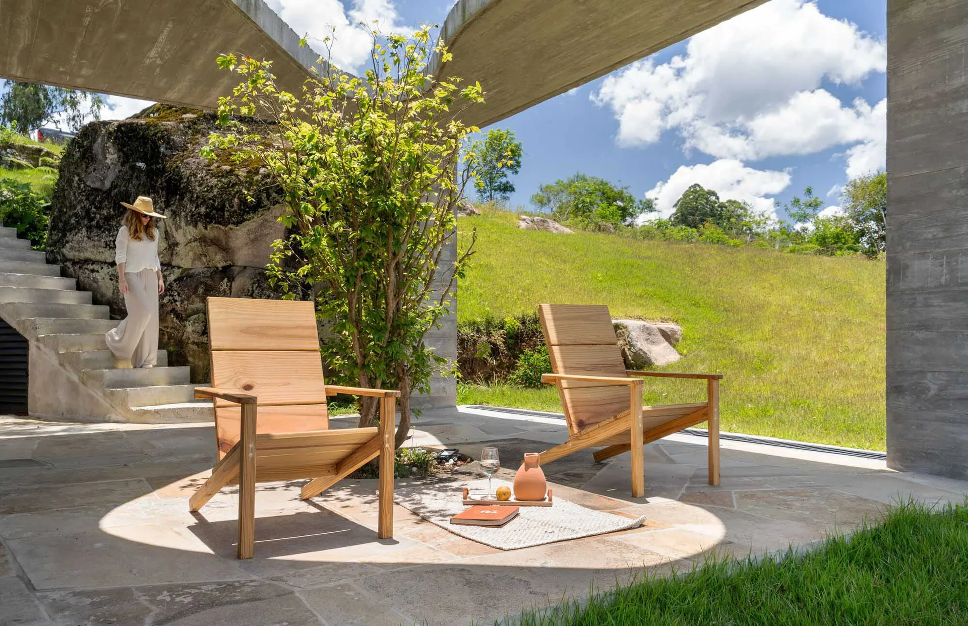 Two wooden Adirondack chairs on a sunny day with concrete walls and a grassy hill in the background.
