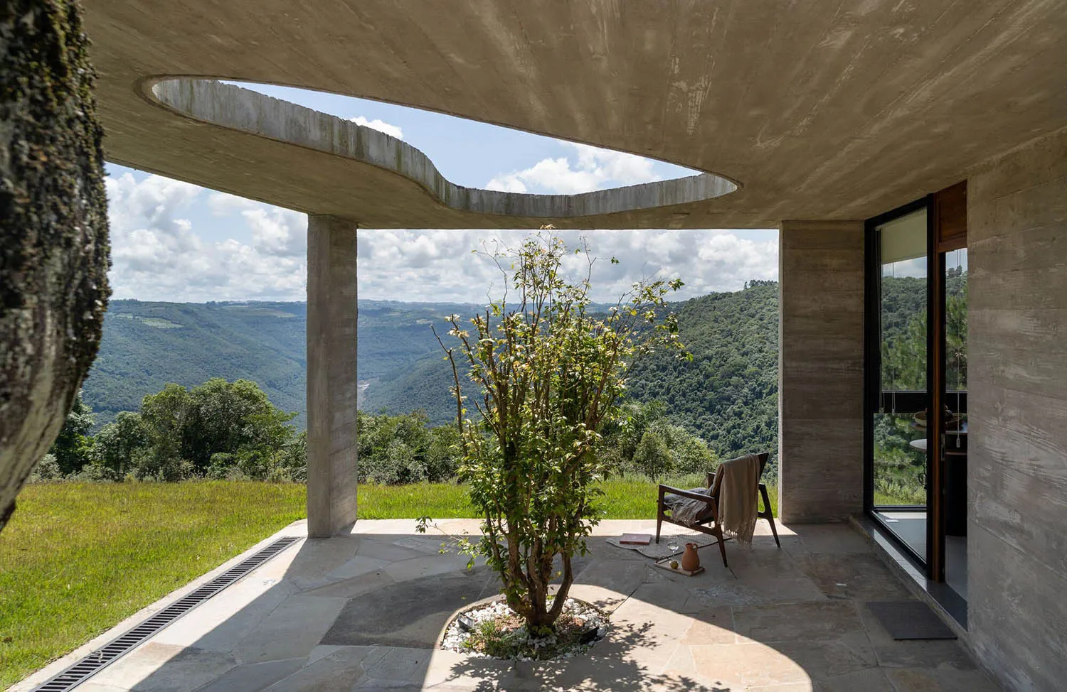 A modern residential interior featuring a concrete ceiling and floor, with a tree and chair on the patio.