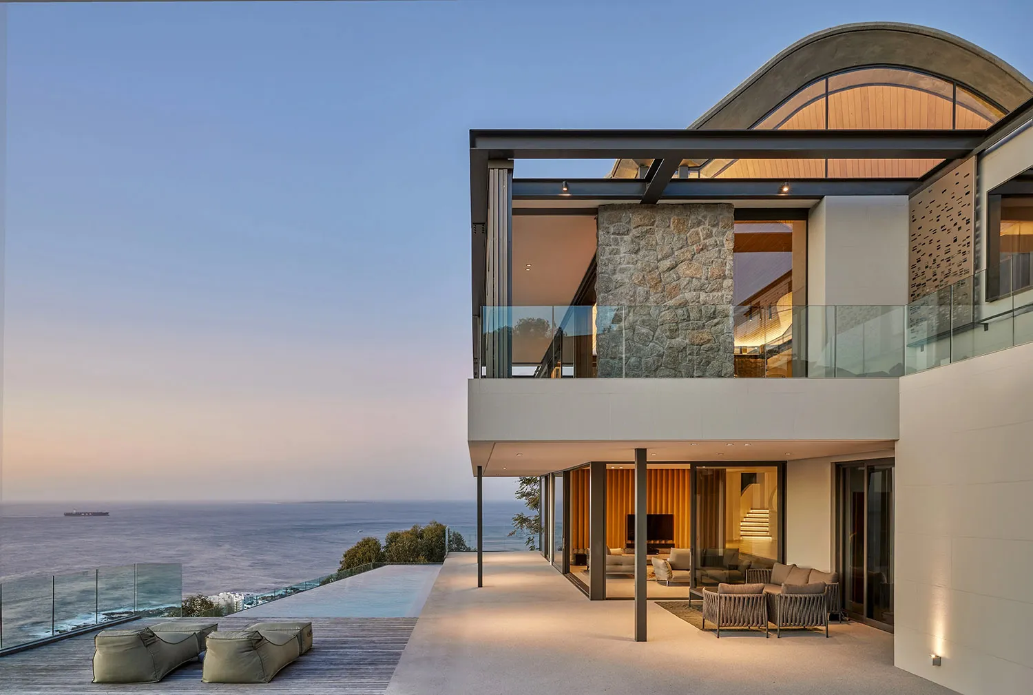 Modern house with concrete exterior and glass walls overlooking the ocean at dusk.