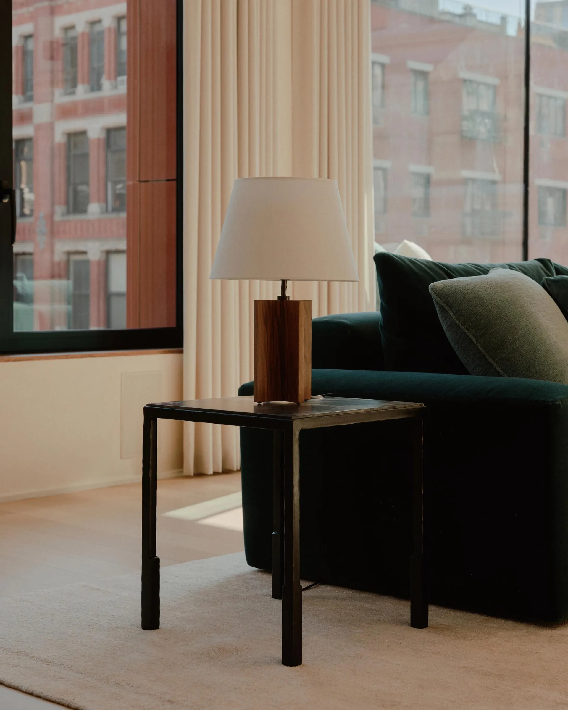 A modern minimalist living room featuring a wooden side table with a white lamp shade, a green velvet sofa, and a large windo