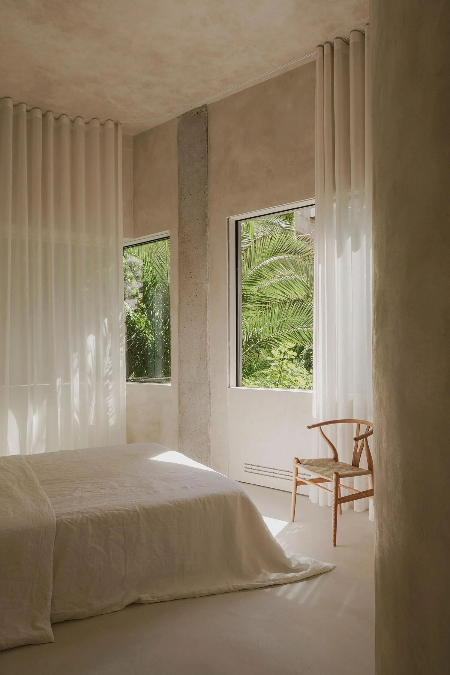 A minimalist bedroom with natural fibers, featuring a bed with white sheets, a wooden chair, and sheer curtains.