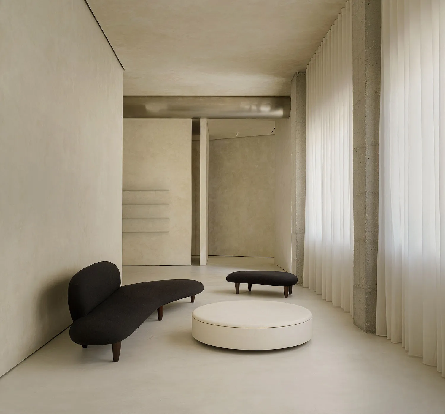 A minimalist living room featuring a black curved sofa, a round white ottoman, and a concrete floor.