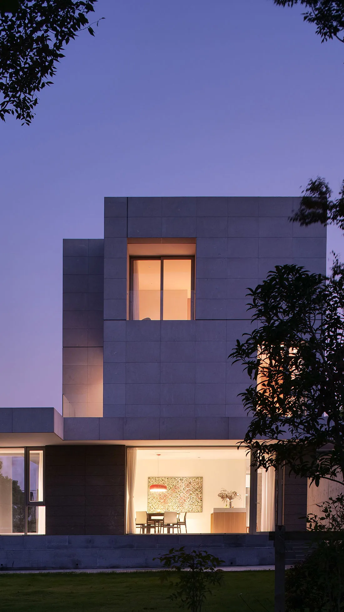 A modern, residential building with concrete exterior at dusk, featuring illuminated windows and a serene garden.