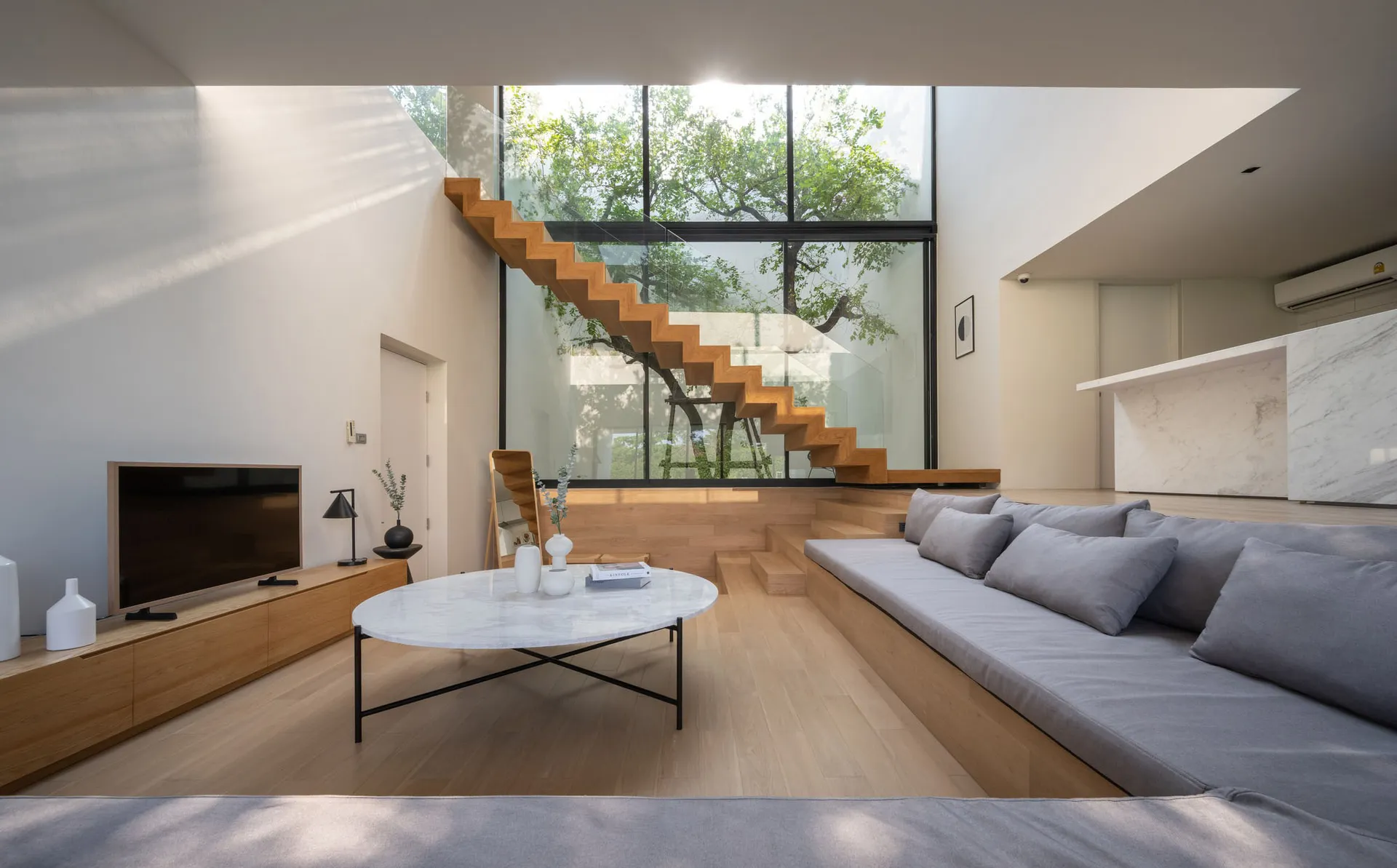 A minimalist living room with wood flooring and a large sofa, featuring a staircase and large window.