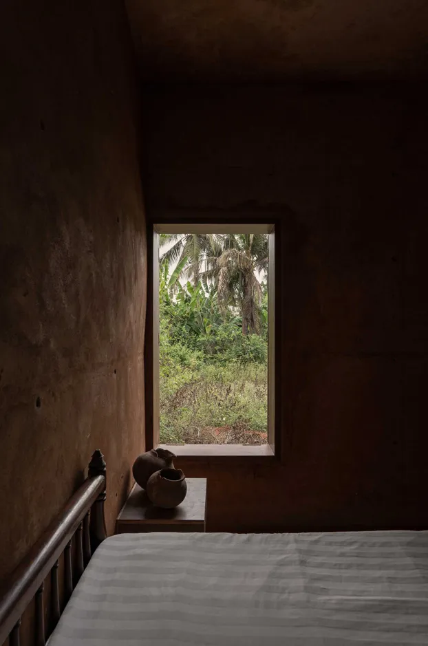 A minimalist bedroom with concrete walls, a bed, and a window overlooking greenery.