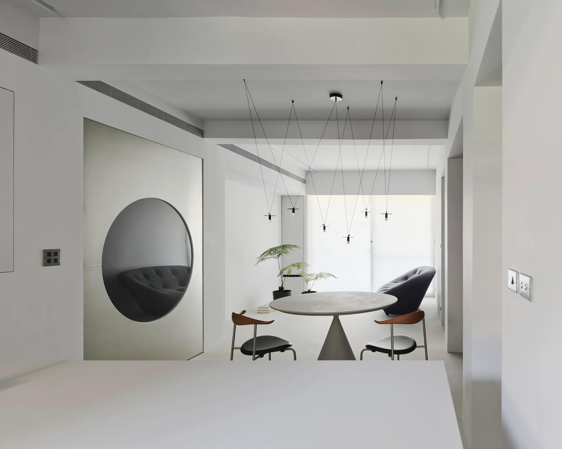 Minimalist dining room with concrete table and chairs, modern hanging lights, and circular mirror on metal panel wall.