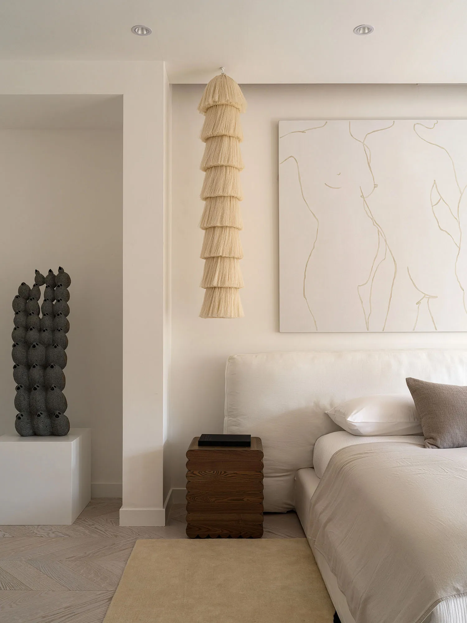 A minimalist bedroom with soft, neutral tones featuring a white bed, wooden nightstand, and fabric art hanging above the bed.