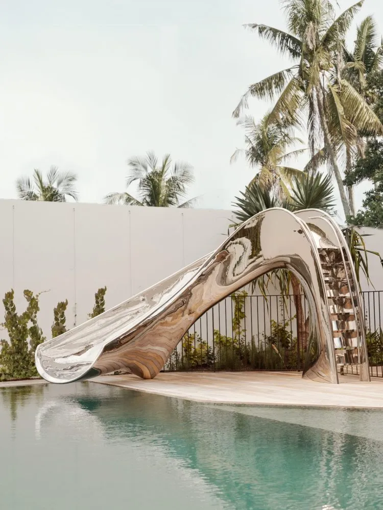 A sleek, metallic outdoor slide leading into a tranquil pool surrounded by lush greenery.