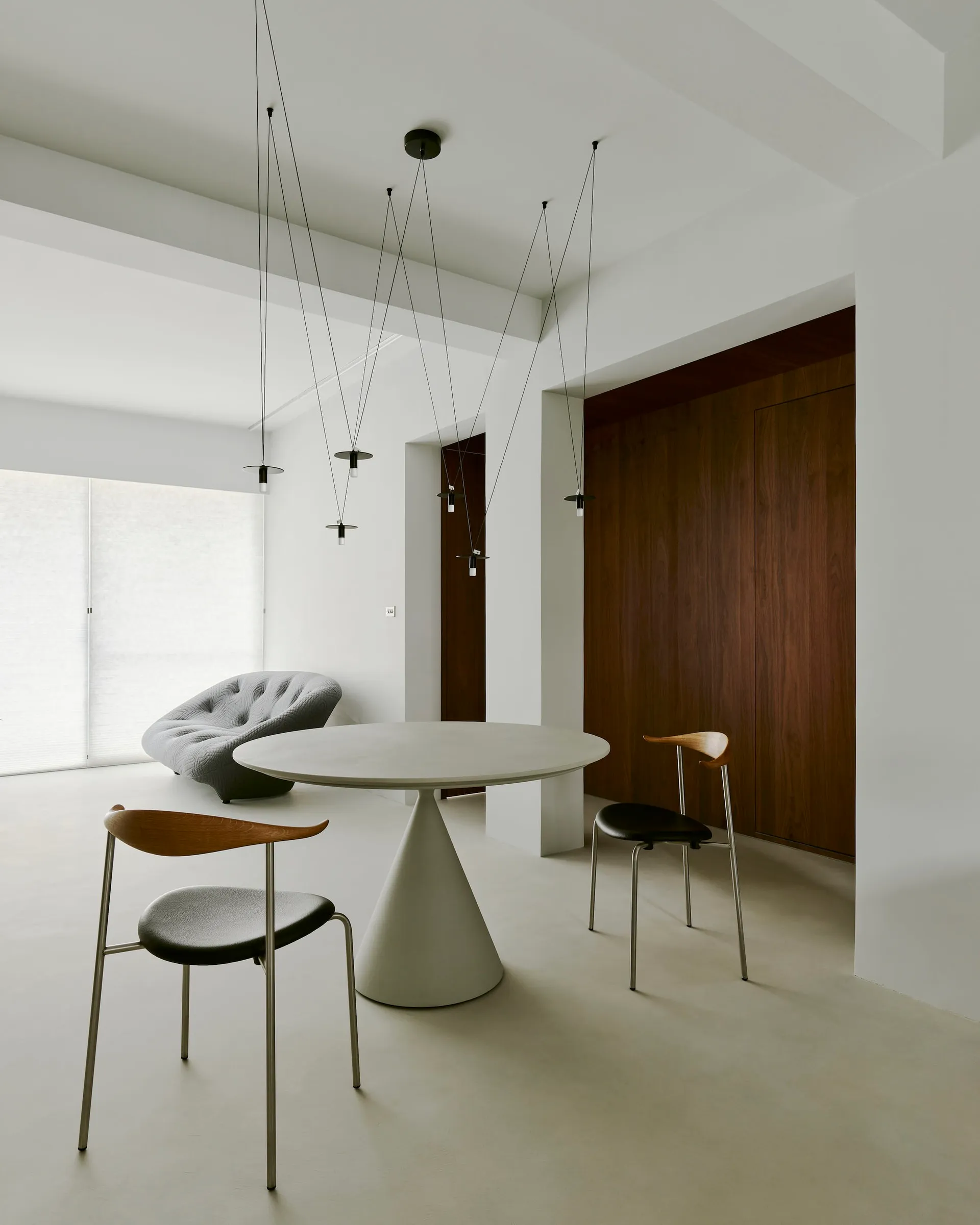 A modern dining room featuring a round, white, cone-shaped table with minimalist black chairs, and a gray tufted armchair in