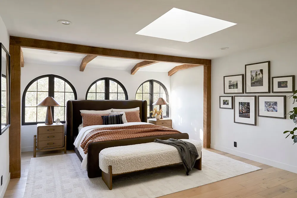 A minimalist bedroom featuring a white wooden floor, a brown leather bed with white and orange bedding, and an arrangement of