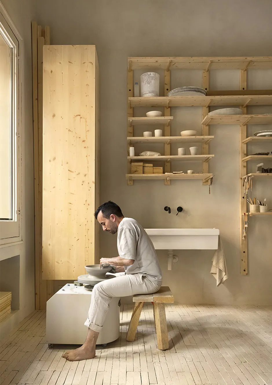 A minimalist pottery studio featuring wooden shelves and a bench, with a man working on a potter's wheel.