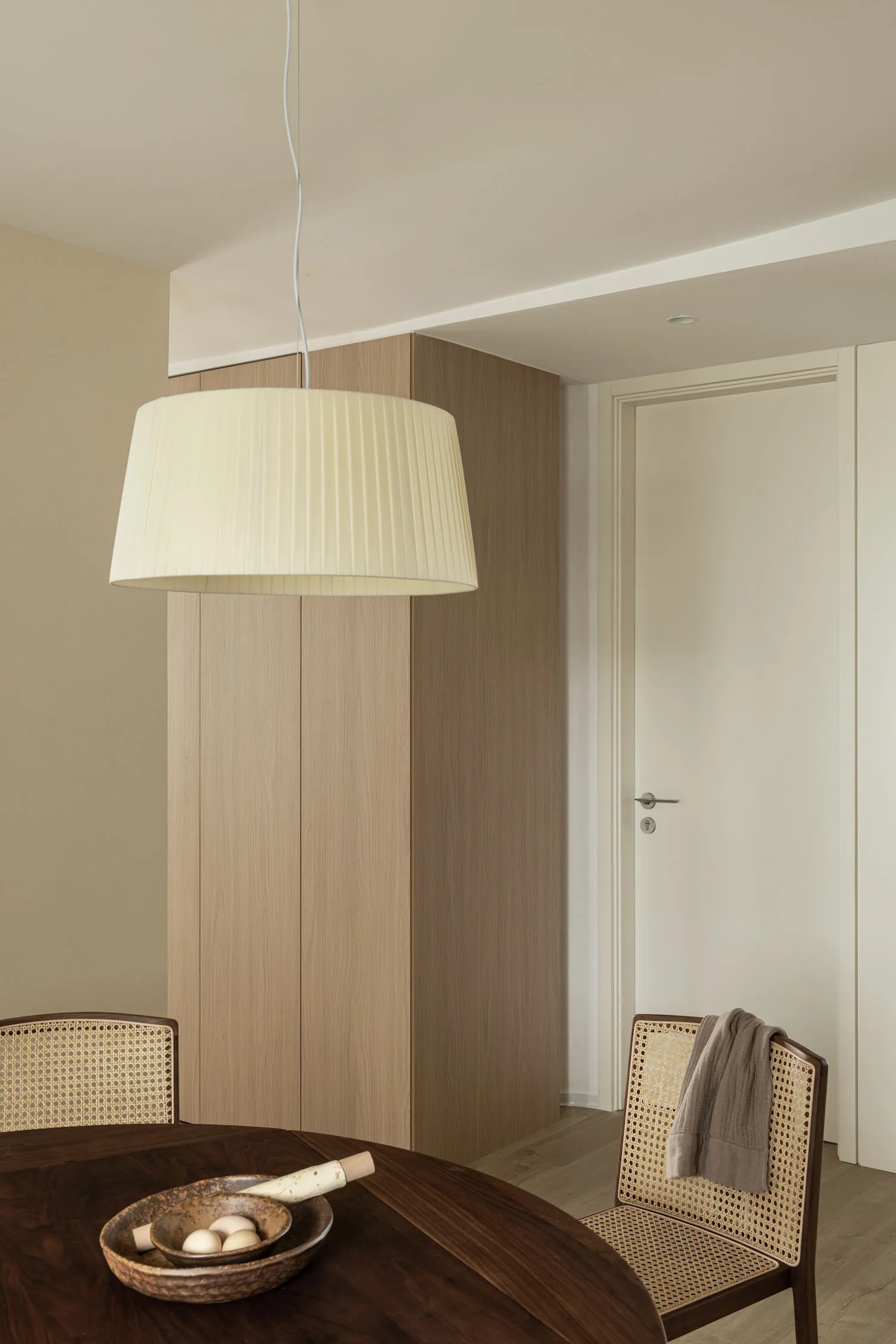 A modern dining room with a wooden table, chairs with woven backs, and a pleated pendant light hanging above.
