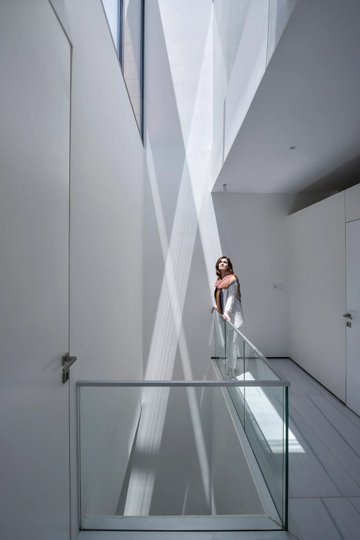 A woman stands on a glass wall railing in a modern, white interior space.