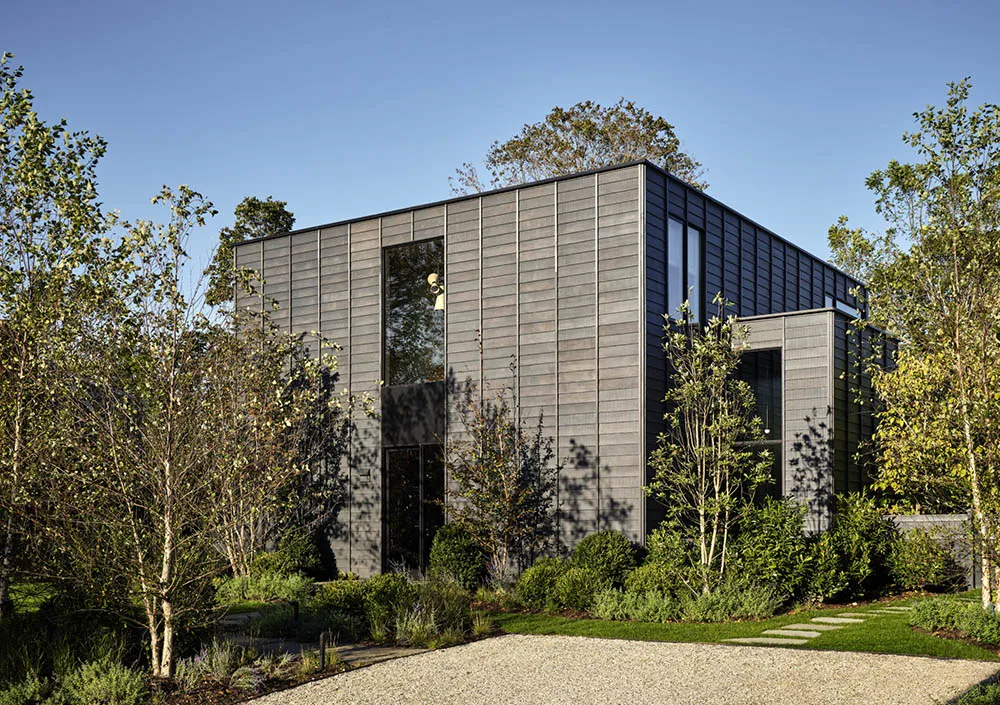 A modern, metal-clad house with large windows and a gravel driveway surrounded by greenery and young trees.