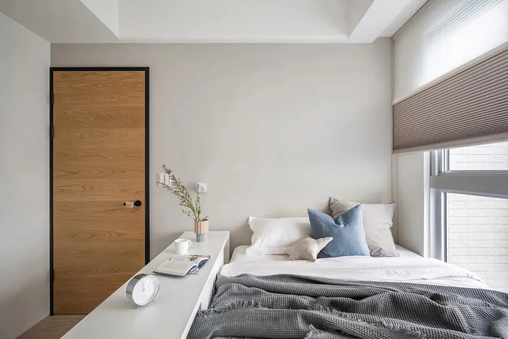 A minimalist bedroom with neutral-toned walls, bedding, and window treatments, featuring a modern wooden door and a sleek whi