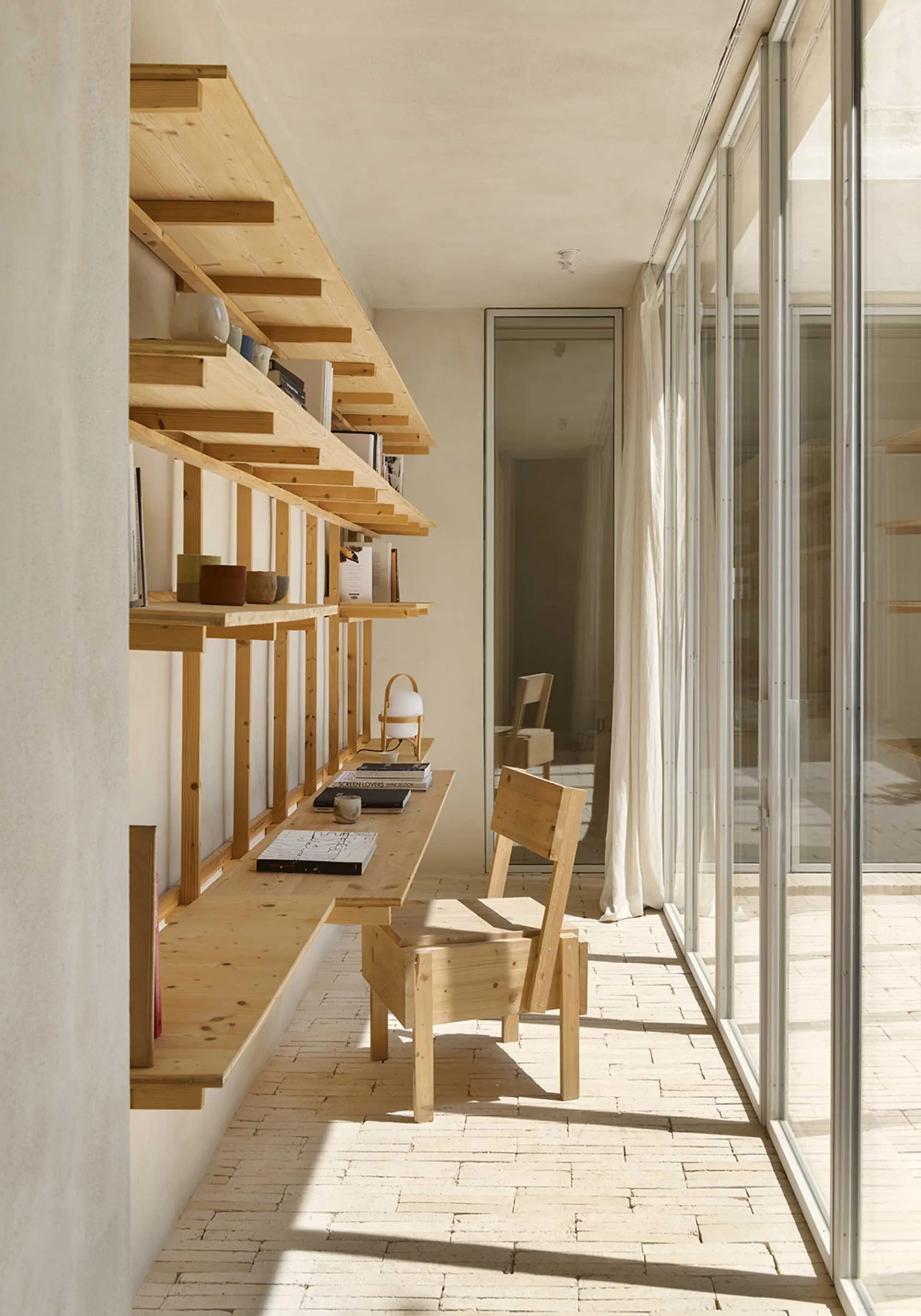A minimalist home office featuring wooden furniture, shelves, and a desk with books and a lamp.