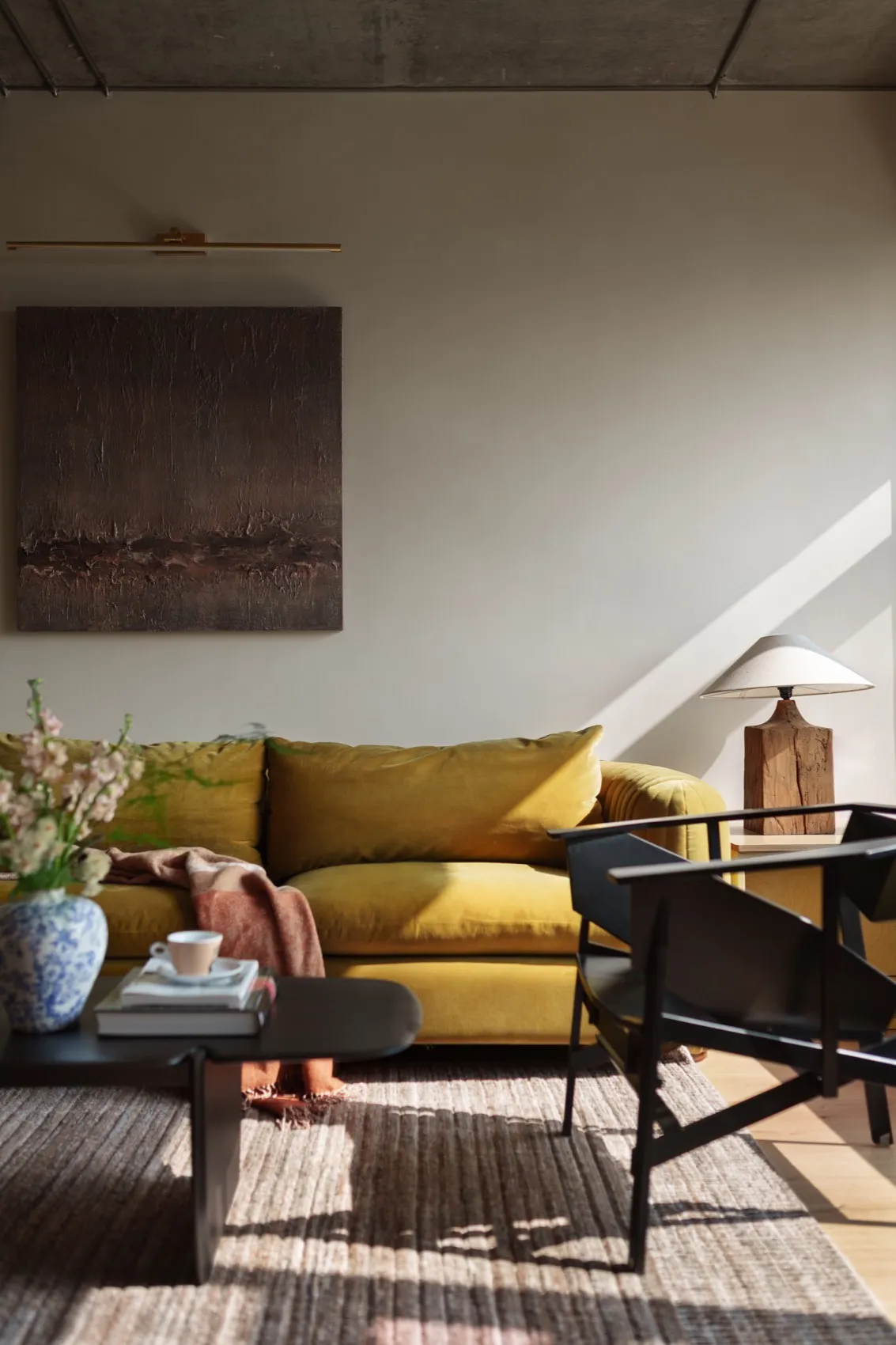 A minimalist living room with a mustard yellow velvet sofa, black chairs, and a wooden lamp on a wooden table with a vase of 