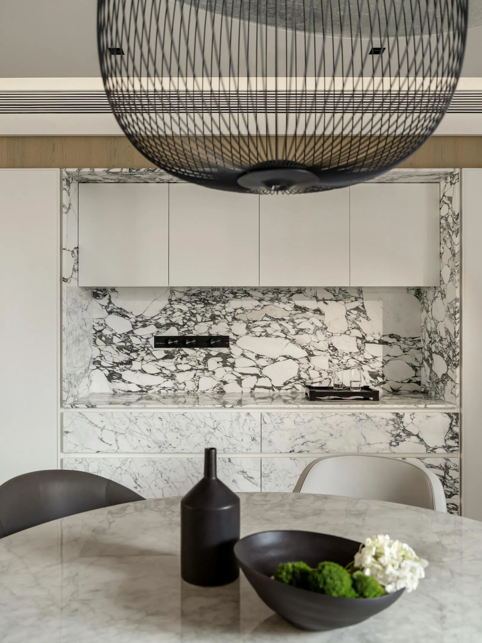 A modern kitchen featuring marble countertops and cabinets against a white marble backsplash, with a sleek vase and bowl of f