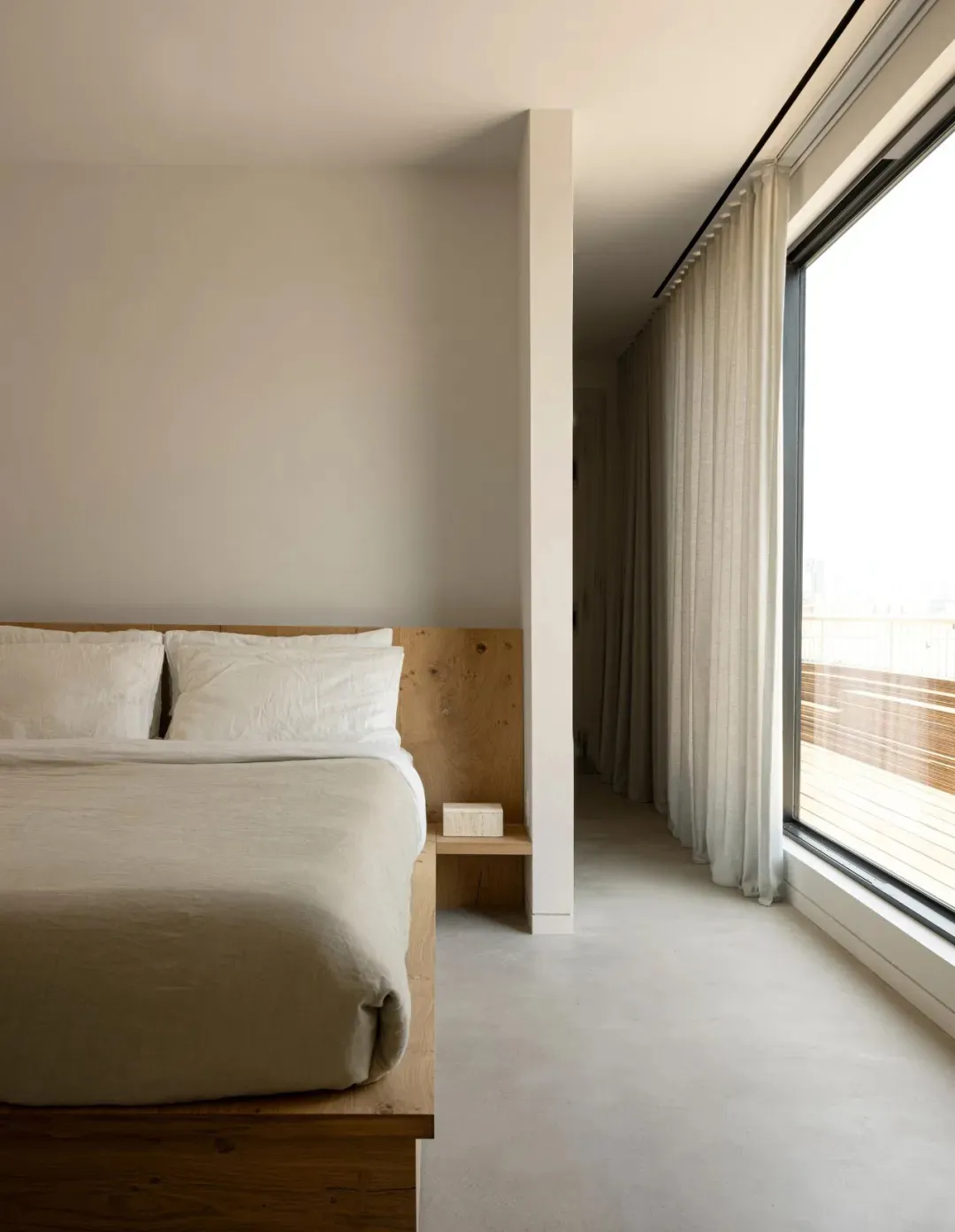 A minimalist bedroom with a wooden bed platform, concrete floor, and large window with sheer curtains.