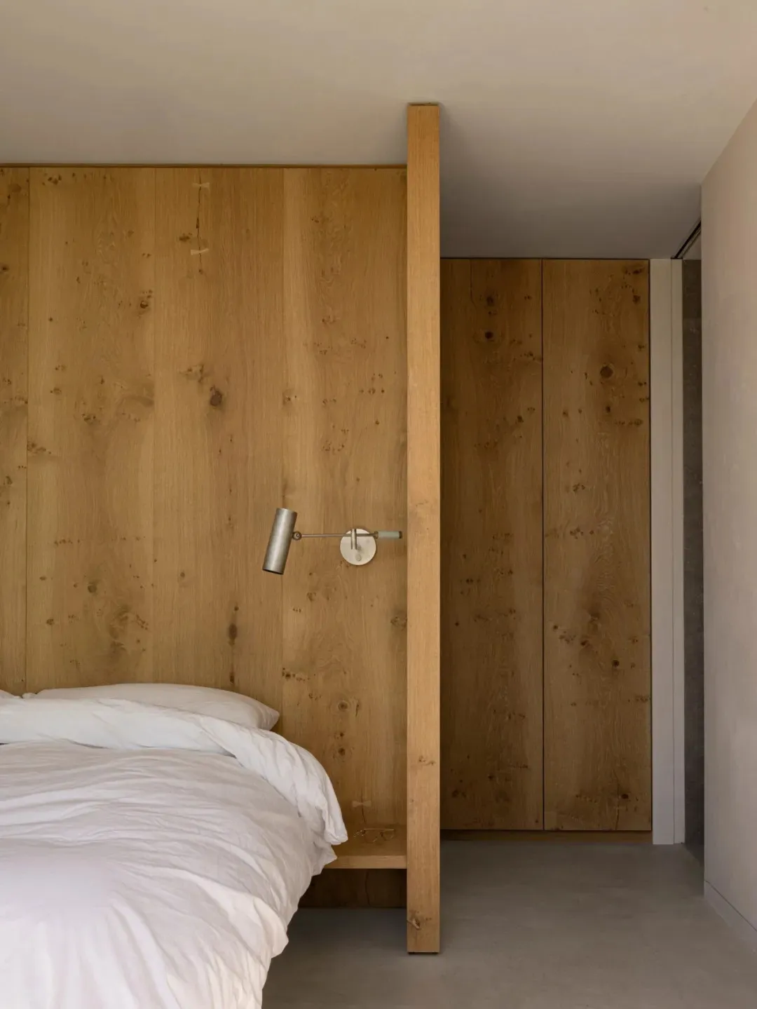 A minimalist bedroom featuring wooden paneled walls, a simple bed with white bedding, and a wall-mounted lamp.