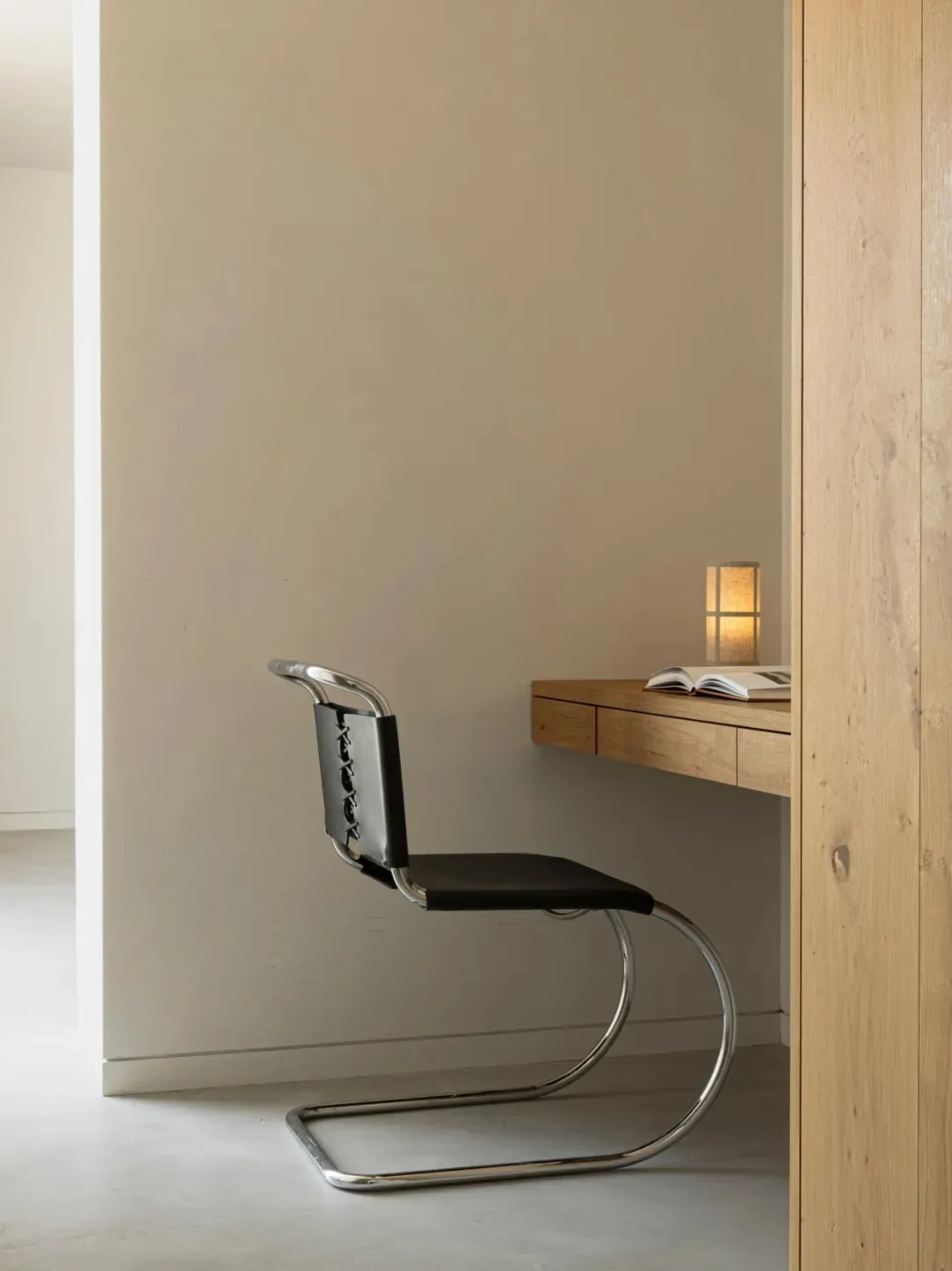 A minimalist black leather chair with chrome frame on a hardwood floor, next to a wooden desk with an open book and a small l