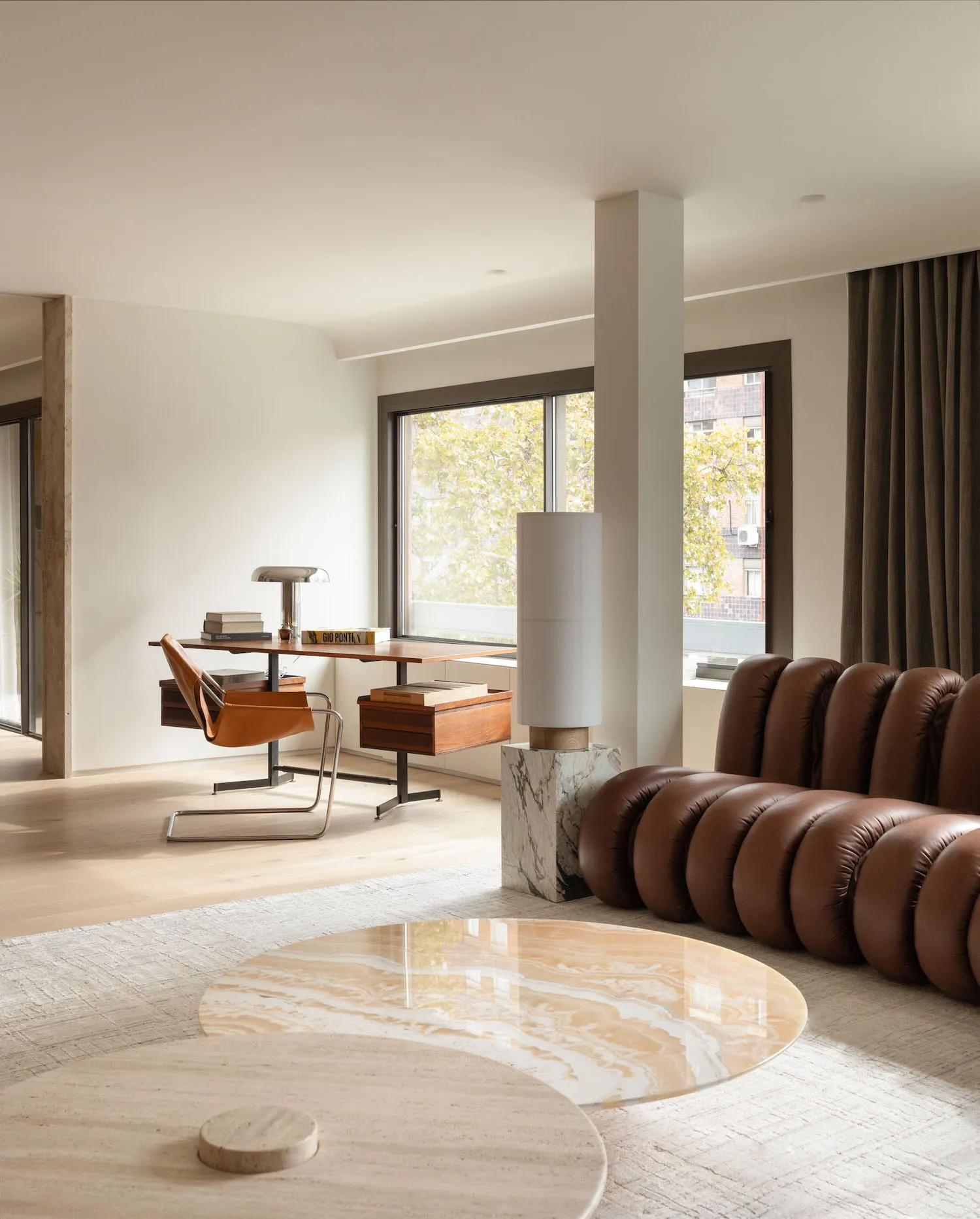 A minimalist living room with a round marble coffee table, brown leather sofa, and wooden desk.