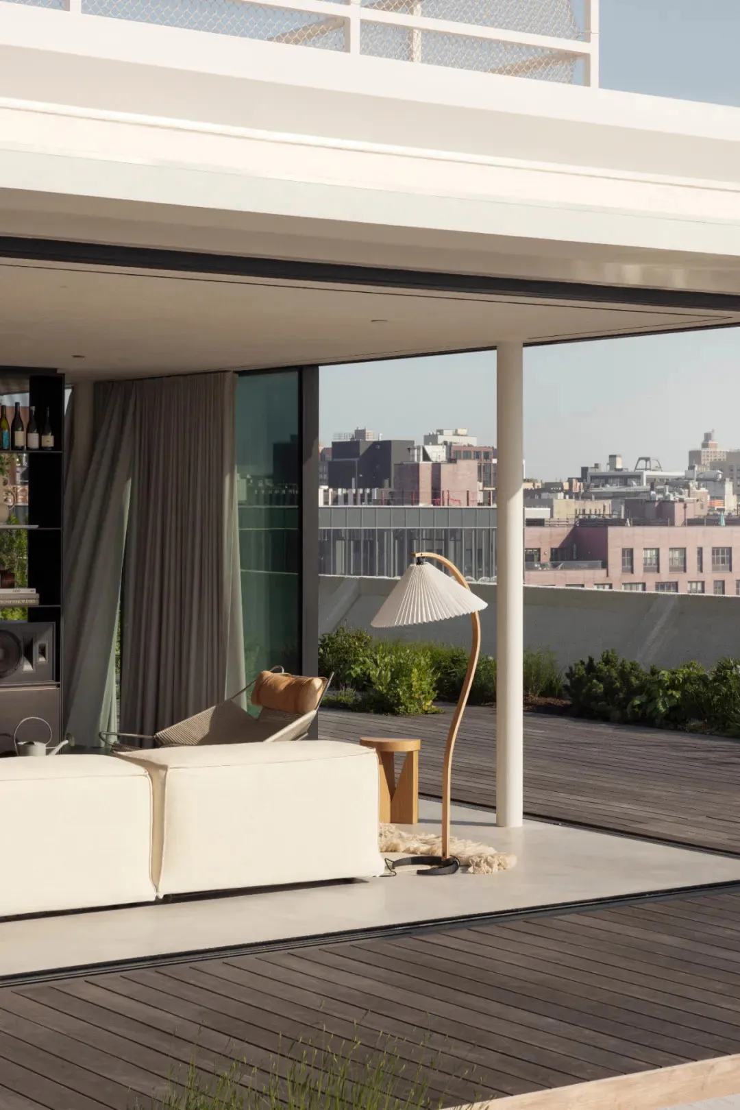 A minimalist living room with a white sofa, a white floor lamp with a curved base, and a wooden deck overlooking a cityscape.