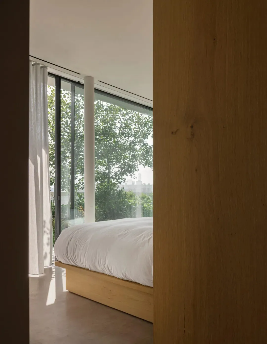 A minimalist bedroom featuring a wooden bed platform and large windows looking out to greenery.